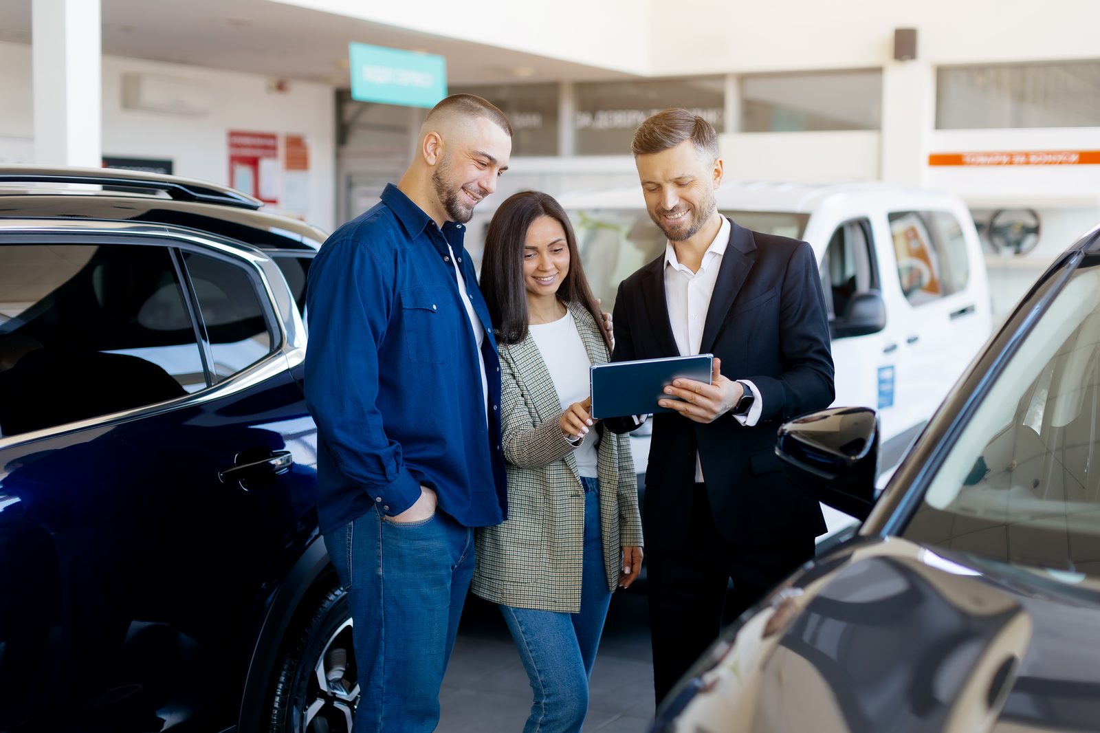 Car salesman showing details on tablet to young couple in modern dealership showroom